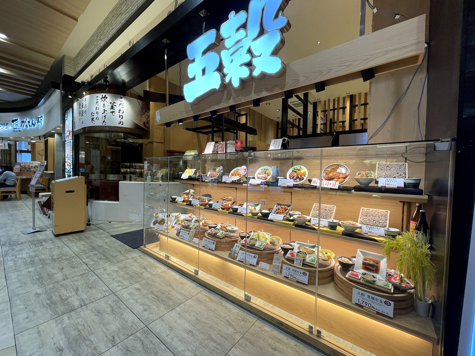 和食処 五穀の外観。青く光る「五穀」の看板と、食品サンプルが並ぶ大きなショーケース