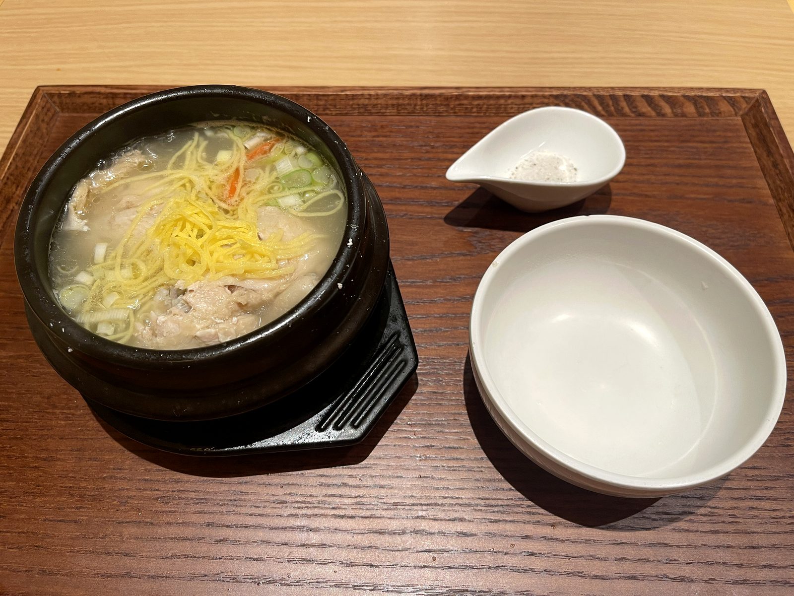 自家製サムゲタンのハーフ。黒い土鍋に丸鶏と錦糸卵、ねぎ、にんじん。脇に塩胡椒の器と取り皿
