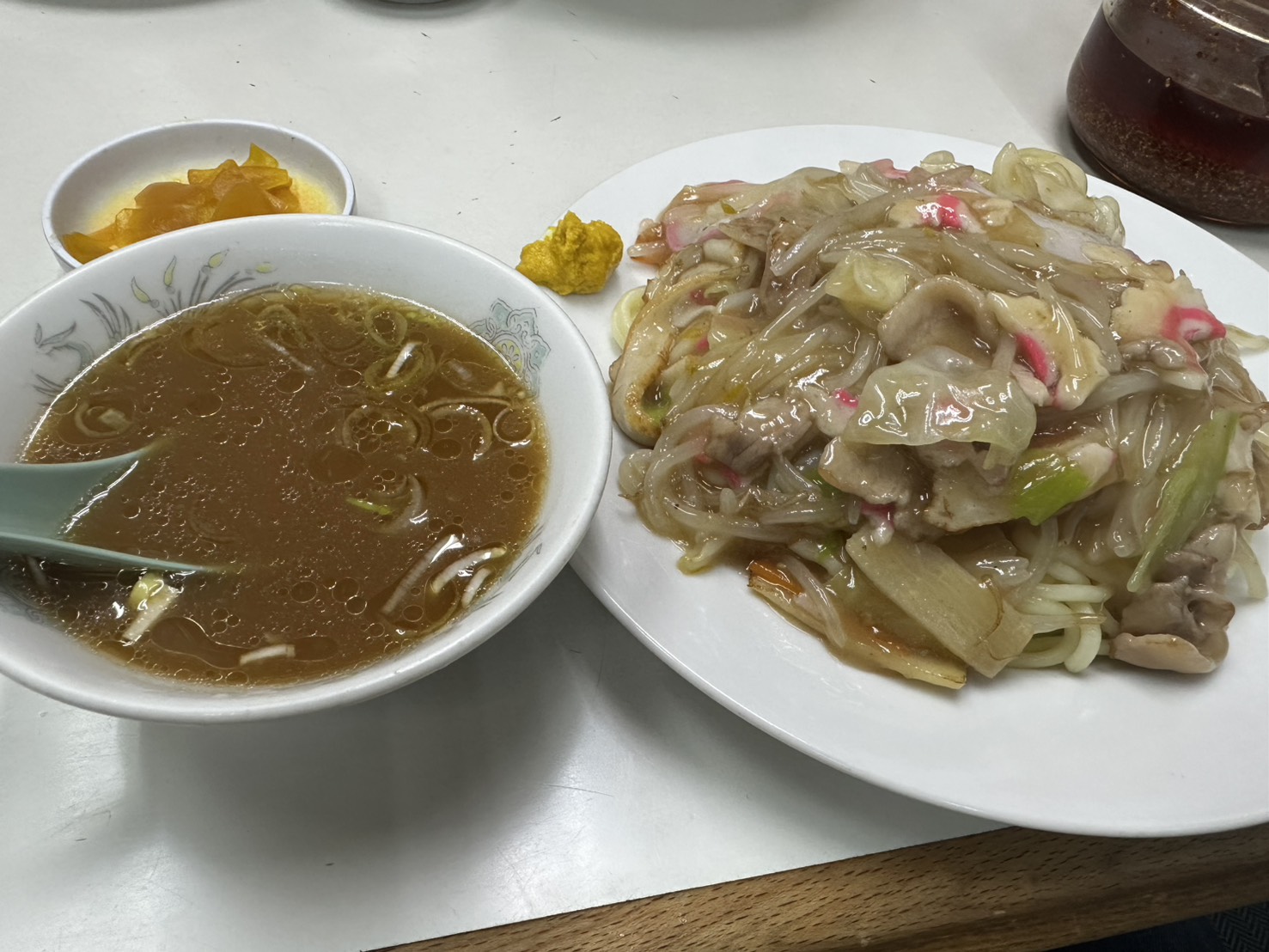 やわらかい太麺にあんかけをかけた皿うどんと、別皿の茶色い醤油スープ、小皿の黄色い沢庵