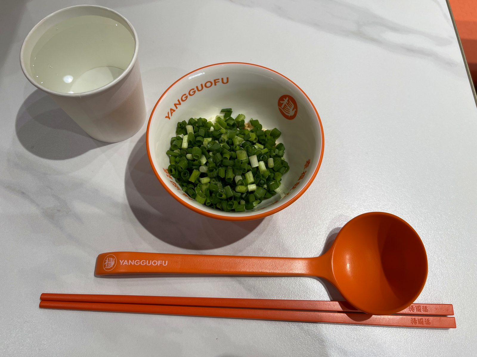 紙コップの水とYANGGUOFUの小鉢に入った刻みネギ、オレンジのレンゲと箸の卓上セット